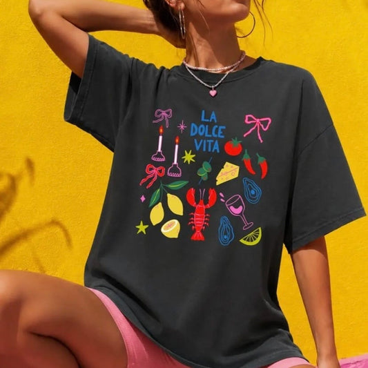 A woman wearing a dark charcoal gray oversized t-shirt with a colorful graphic print that reads 'LA DOLCE VITA' surrounded by summer-themed illustrations, posed against a bright yellow wall.
