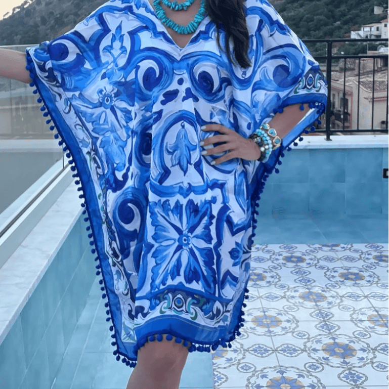 A woman wearing a blue and white patterned kaftan dress with pom-pom trim, standing on a tiled terrace with a mountainous landscape in the background.
