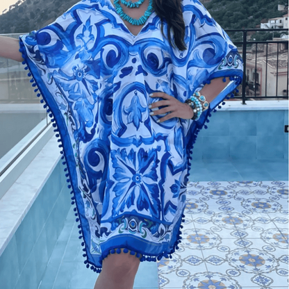 A woman wearing a blue and white patterned kaftan dress with pom-pom trim, standing on a tiled terrace with a mountainous landscape in the background.