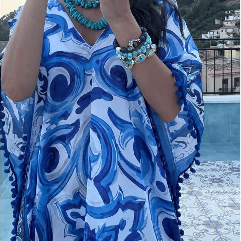 A woman takes a mirror selfie wearing a blue and white patterned kaftan dress with turquoise jewelry, standing outdoors near a pool and hills.