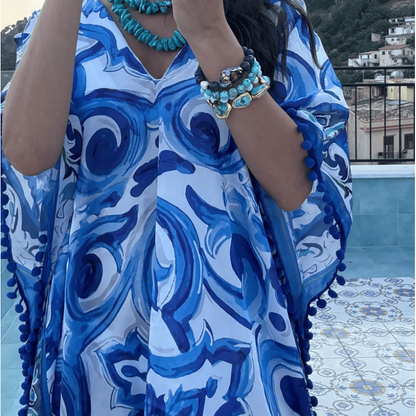 A woman takes a mirror selfie wearing a blue and white patterned kaftan dress with turquoise jewelry, standing outdoors near a pool and hills.
