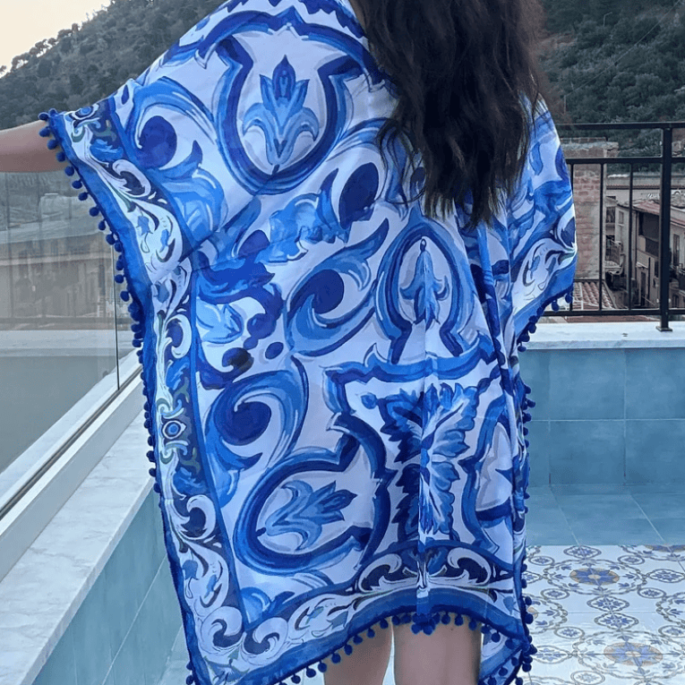 A woman wearing a blue and white patterned kaftan cover-up with pom-pom trim, photographed outdoors on a balcony with a pool and hills in the background.
