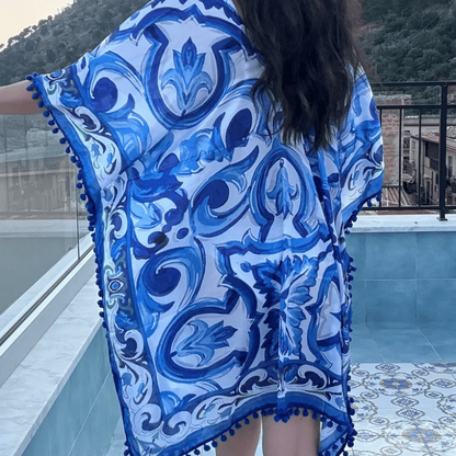 A woman wearing a blue and white patterned kaftan cover-up with pom-pom trim, photographed outdoors on a balcony with a pool and hills in the background.