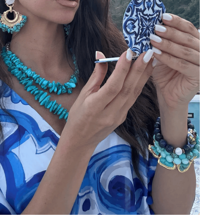 A woman outdoors holds a small blue-and-white patterned tile or mirror while wearing turquoise jewelry and a blue-and-white patterned top. The photo appears to be a social media or website screenshot with a promotional banner at the bottom.